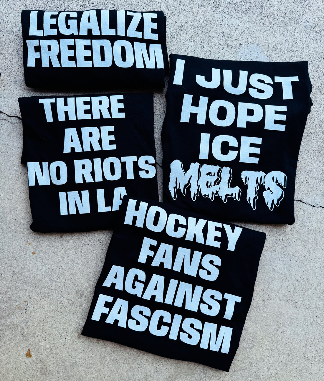 four tee shirts laying on the concrete. they read from top to bottom ,left to right: legalize freedom; there are no riots in LA; I just hope the ICE melts; hockey fans against fascism. tee shirts are black with white bold text